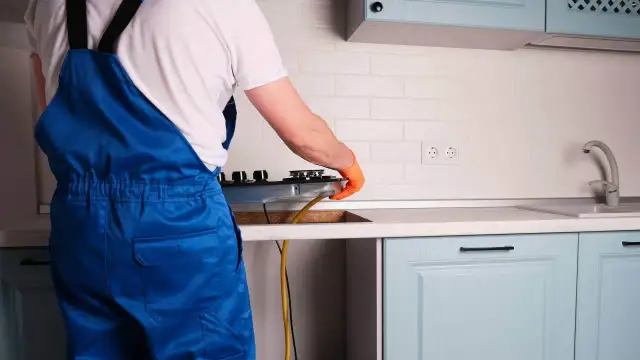 Instalator w niebieskim kombinezonie podłącza kuchenkę gazową w kuchni.