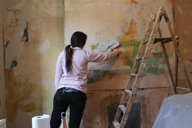 Kobieta z pędzlem w ręku usuwa tynk strukturalny ze ściany. Obok stoi drabina.