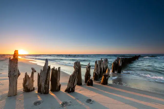 Zachód słońca nad Bałtykiem, z drewnianymi słupkami na plaży i falami.