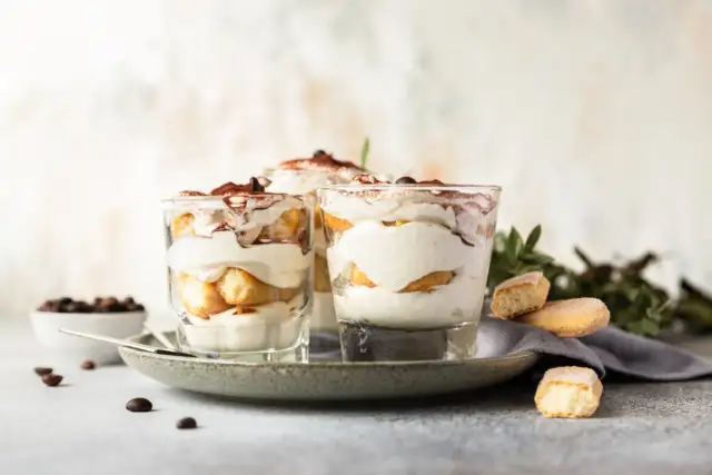 Pyszne desery włoskie: tiramisu w szklankach, udekorowane kakao i kawą. Obok kawałki biszkoptów i ziarna kawy.