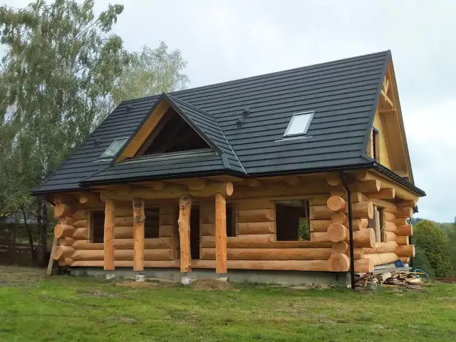 Dom z bali w trakcie budowy, z widocznymi belkami, oknami i czarnym dachem.