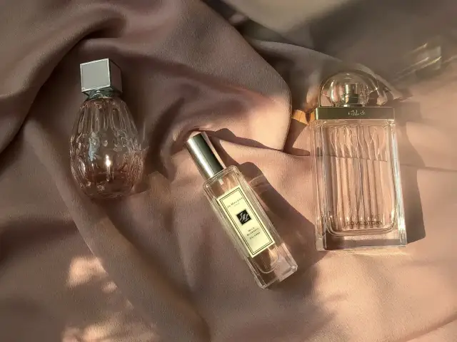 Drei Parfümflaschen auf rosafarbenem Stoff, im Sonnenlicht.