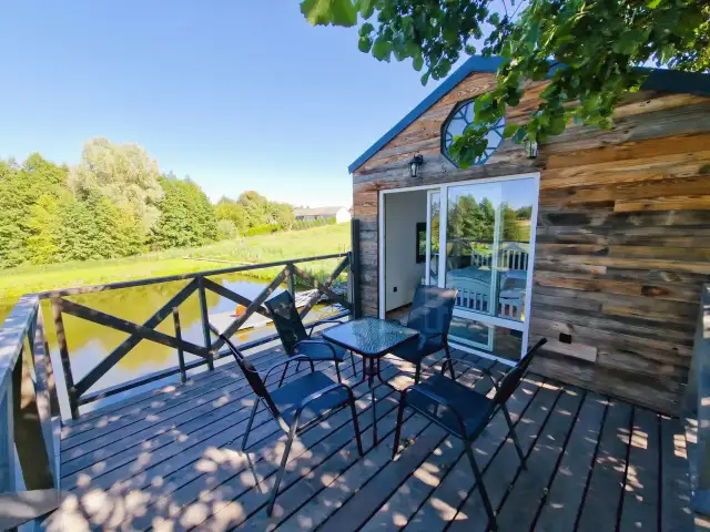 Drewniany domek nad jeziorem z tarasem, stołem i krzesłami.