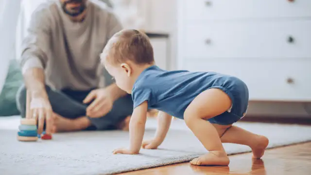 Dziecko w niebieskim body raczkuje na dywanie, obok siedzi ojciec z zabawkami.