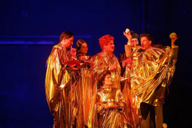 Aktorzy w złotych szatach, trzymający filiżanki i książki, tworzą scenę z najlepszych spektakli teatralnych.