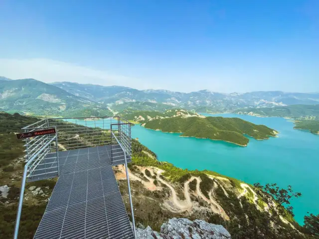 Widok z platformy widokowej na turkusowe wody jeziora Bovilla, otoczonego zielonymi wzgórzami i górami.