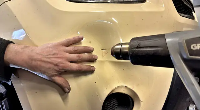 Dłoń przytrzymuje wgniecenie na zderzaku, podczas gdy opalarka jest używana, by wyprostować odkształcony plastik.