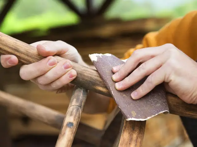 Dłonie szlifują drewniany mebel papierem ściernym, dbając o odpowiednią gradację.