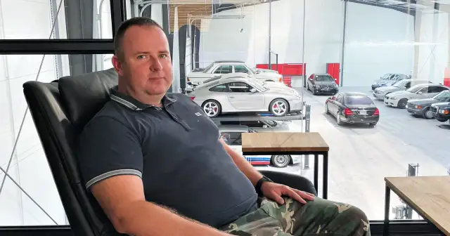 Robert Michalski, w wieku około 40-50 lat, siedzi w czarnym fotelu. W tle widać kolekcję samochodów.