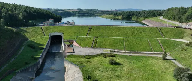 Zapora wodna z budynkiem elektrowni i kanałem odpływowym na tle jeziora i lasów.