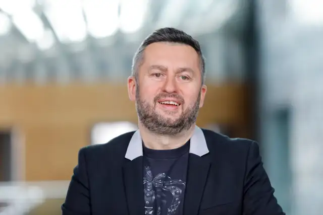 Uśmiechnięty mężczyzna w czarnej marynarce i t-shircie z nadrukiem. Ciekawe, ile zarabia Norbi w Kole Fortuny?