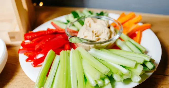 Talerz z pokrojonymi w słupki warzywami: marchewką, ogórkiem, papryką i selerem, z miseczką hummusu.