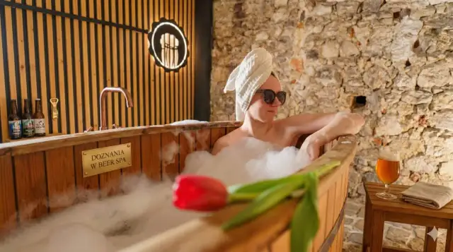 Relaks w beer spa w Zakopanem. Kobieta w wannie z pianą, ręcznikiem na głowie i okularach przeciwsłonecznych, obok piwo i tulipan.