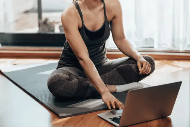 Kobieta w stroju sportowym siedzi na macie, korzystając z laptopa. Czy można ćwiczyć codziennie? Tak, jeśli masz dostęp do inspiracji online!