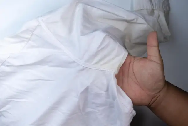 Dłoń pokazuje białą koszulę z żółtymi, twardymi plamami po antyperspirancie pod pachą.
