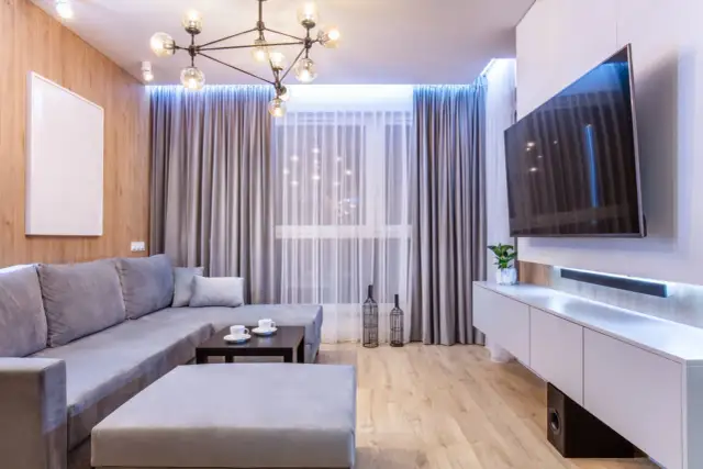 Nowoczesny salon w małym mieszkaniu 40 m: szara sofa, stolik kawowy, TV na ścianie i drewniana ściana.