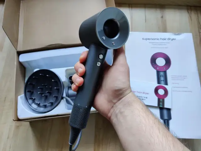 Dłoń trzymająca czarną suszarkę do włosów Dyson Supersonic obok pudełka i dyfuzora.
