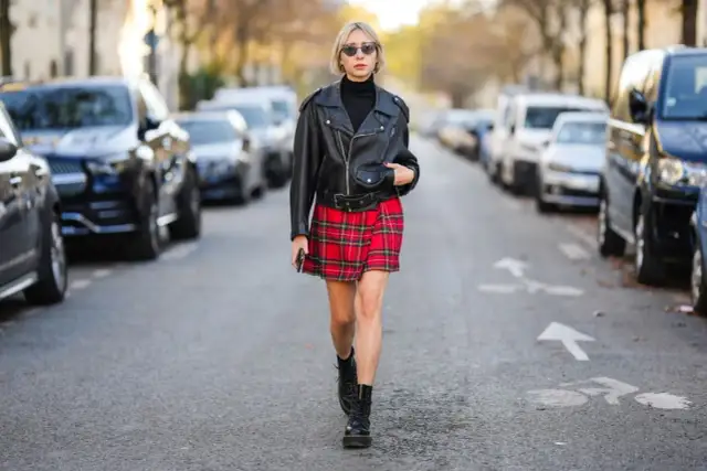 Kobieta w skórzanej ramonesce, czarnym golfie i czerwonej spódnicy w kratę prezentuje styl ubioru grunge. Na nogach ma ciężkie buty.