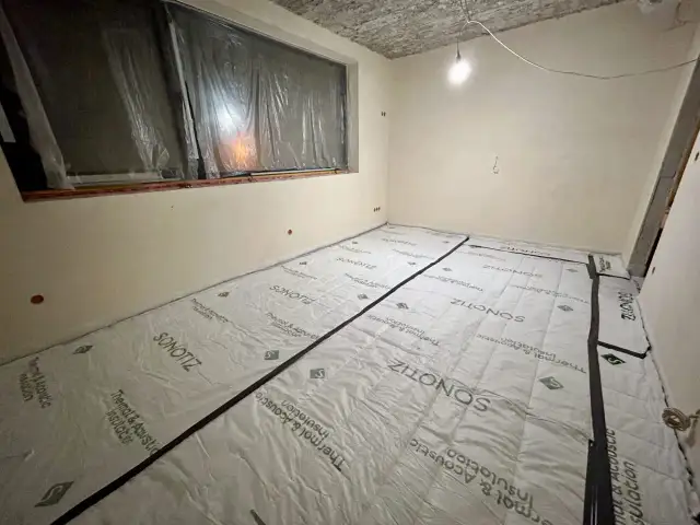 W trakcie prac remontowych w bloku, podłoga pokryta jest materiałem do wyciszenia. Widać okno zabezpieczone folią i sufit z widocznymi instalacjami.