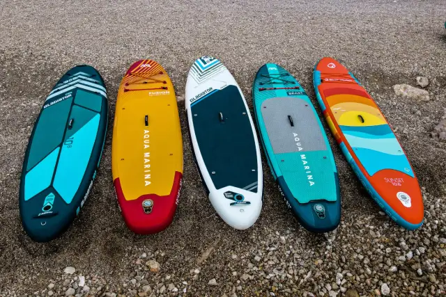 Pięć desek SUP w różnych kolorach i wzorach leży na kamienistej plaży.