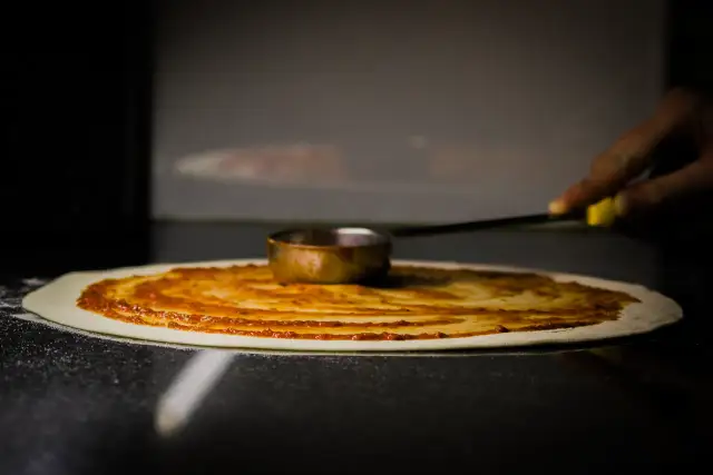 Ręka rozprowadza sos pomidorowy na cieście, tworząc idealny zaczyn na pizzę.