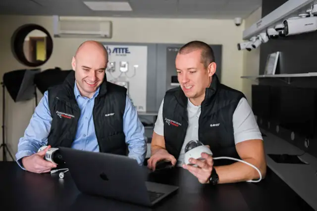 Dwóch mężczyzn omawia systemy monitoringu. Jeden trzyma kamerę, drugi patrzy na laptopa, planując zdalny monitoring.