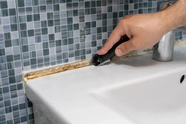 Dłoń z nożykiem usuwa zaschnięty, brudny silikon z białej umywalki przy kafelkach.