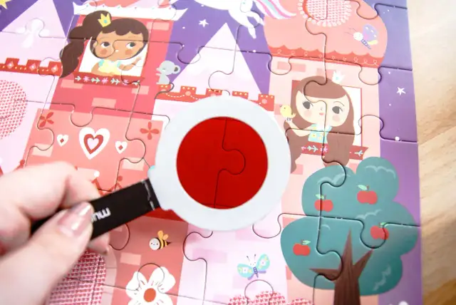 Kolorowa układanka dla dzieci z księżniczkami i zamkiem. Dłoń trzyma lupę, przez którą widać dwa czerwone elementy układanki.