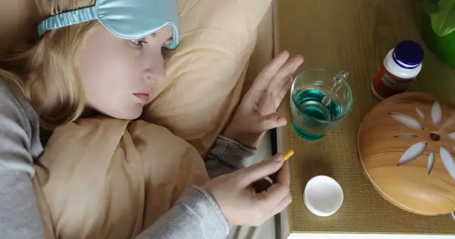 Kobieta w opasce na oczy trzyma kapsułkę, zastanawiając się, melatonina co to. Obok szklanka wody i suplementy.