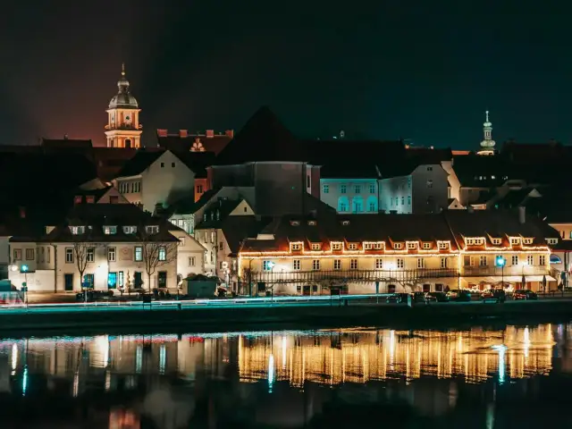 Nocny widok na zabytkowe budynki w Mariborze, Słowenia, z odbiciami świateł w rzece.