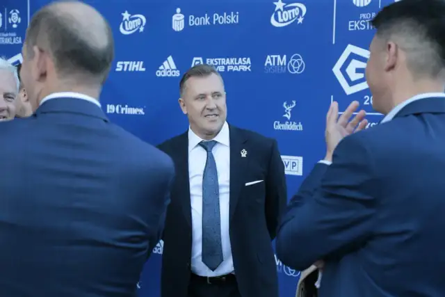 Mężczyzna w garniturze uśmiecha się, stojąc na tle niebieskiej ściany z logotypami sponsorów, być może rozmawiając o tym, ile dostają kluby ekstraklasy.