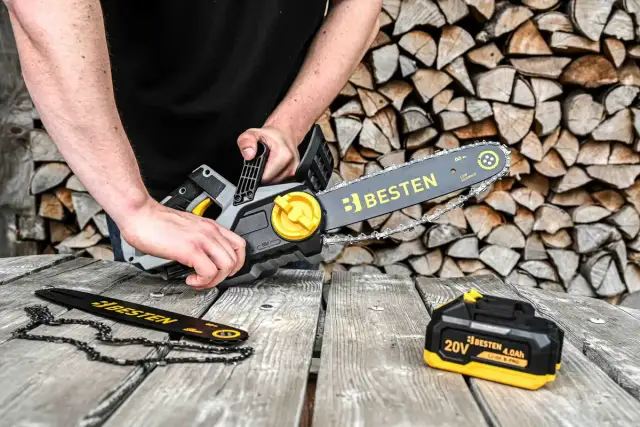 Ręce trzymają piłę łańcuchową Besten, przygotowując się do cięcia drewna. Obok leży zapasowy łańcuch i bateria.