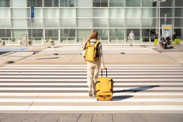 Kobieta z żółtym plecakiem i walizką na kółkach przechodzi przez pasy przed nowoczesnym budynkiem.