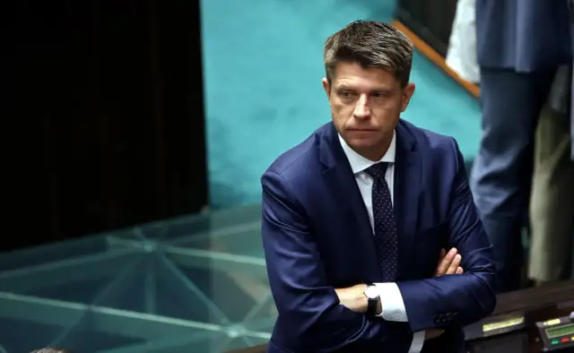 Ryszard Petru w garniturze z rękami skrzyżowanymi na piersi, w tle niebieskie siedzenia.