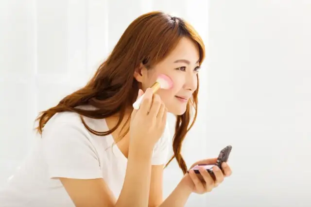 Kobieta nakłada róż na policzki pędzlem, trzymając w dłoni kosmetyczkę.