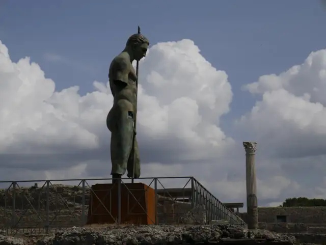 Brązowy posąg mężczyzny z włócznią na tle nieba z chmurami i ruinami Pompejów.