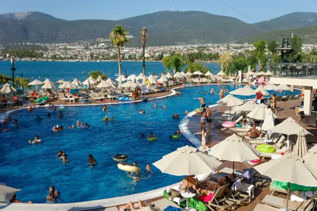 Ludzie kąpią się w basenie, opalają na leżakach pod parasolami. Widok na morze i miasto. Idealne miejsce na wakacje ultra all inclusive.