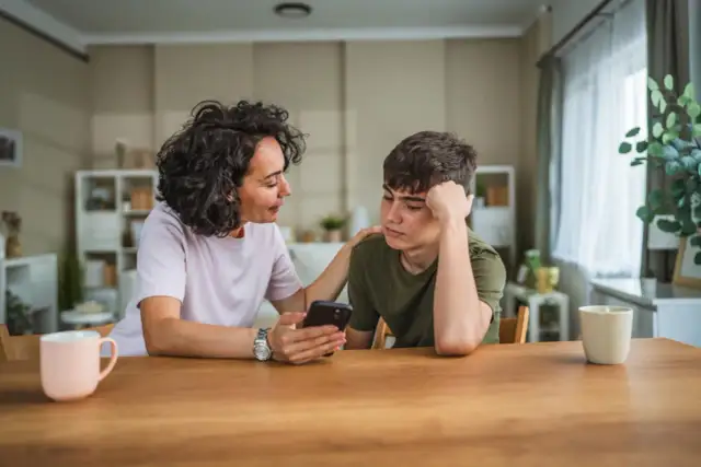 Matka z troską patrzy na telefon syna, zastanawiając się, czy rodzic ma prawo sprawdzać dziecku telefon.