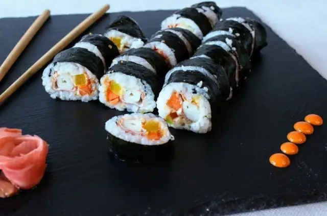 Roladki sushi z surimi, ryżem i warzywami na czarnej desce, obok pałeczki i różowy imbir.