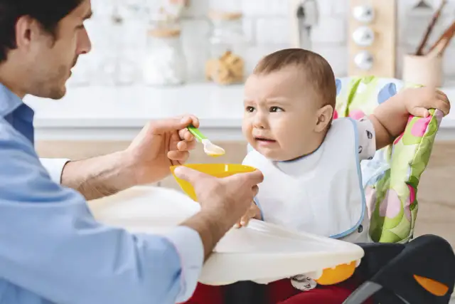 Ojciec karmi dziecko, które nie chce jeść. Czy niemowlę może jeść wątróbkę? Dziecko ma minę wyrażającą niechęć.
