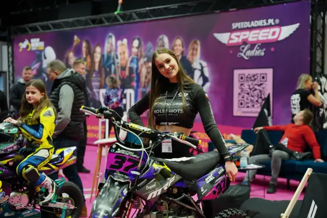 Uśmiechnięta dziewczyna na tle banera z napisem "Warsaw Motorcycle Show" i motocyklem. Obok młoda zawodniczka w żółtym kombinezonie.
