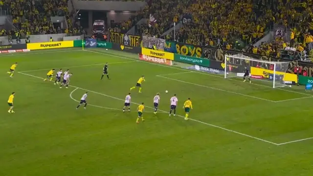 Piłkarze w żółtych i różowych strojach grają w piłkę nożną na boisku, zbliżając się do bramki.