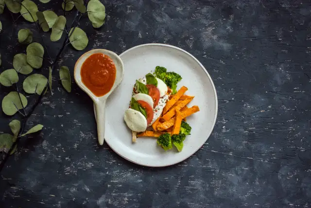 Talerz z batatami, brokułami, mozzarellą i pomidorem, obok łyżka z sosem.