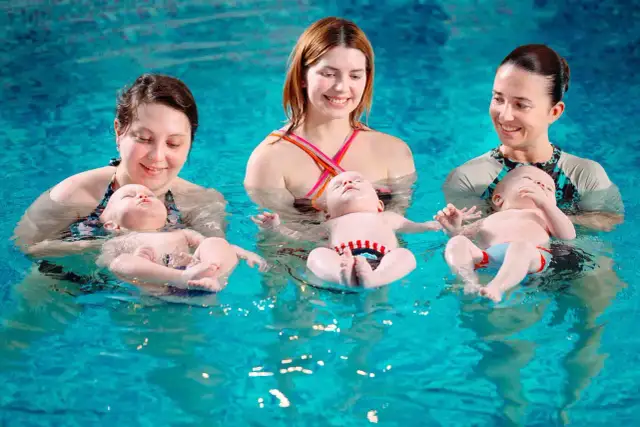 Trzy mamy i ich dzieci w wodzie, każde dziecko w swoim kółku do pływania, uśmiechnięte i zrelaksowane.