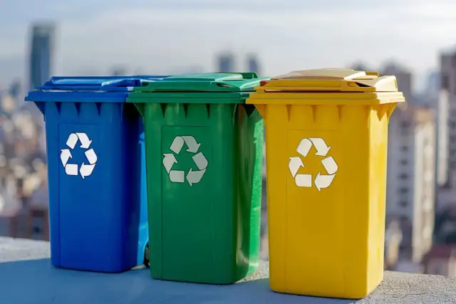 Trzy kolorowe kosze na śmieci: niebieski, zielony i żółty, z symbolami recyklingu. Uczą, jak segregować śmieci dla lepszej przyszłości.