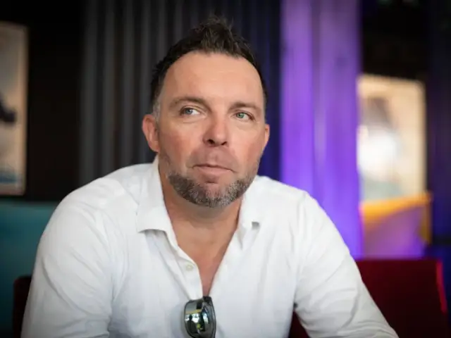 Marcin Wójcik, znany z kabaretu, w białej koszuli, z charakterystyczną fryzurą i zarostem, spogląda w bok.