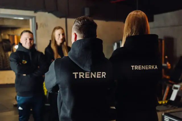 Trzech trenerów w czarnych bluzach z napisami "TRENER" i "TRENERKA" stoi w siłowni.