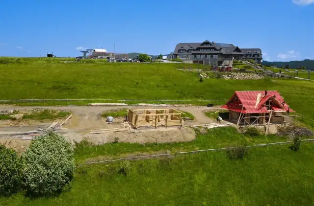 Budowa domu z bali w górach. Widać fundamenty i ściany z bali, a także dom z czerwonym dachem w trakcie prac. To początek budowy, jak wyczyścić dom z bali, by służył latami.
