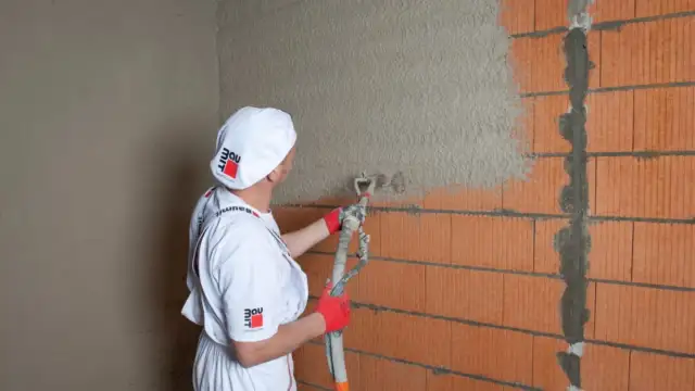 Pracownik w białym ubraniu i czapce z logo Baumit nakłada tynk agregatem na ścianę z pustaków. Obliczasz ile kg tynku na m2?