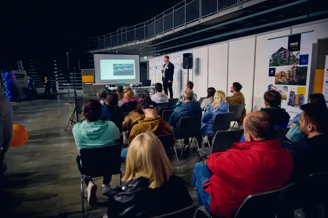 Prezentacja na targach budowlanych. Mężczyzna przemawia do zgromadzonej publiczności, która siedzi na krzesłach.